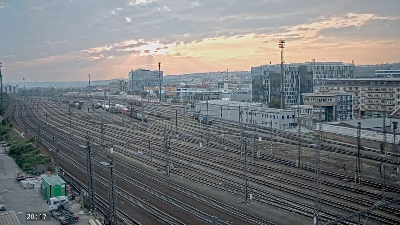 Numerous train tracks and railway cars extend into the distance beneath a partly cloudy sunset sky, next to buildings and structures.