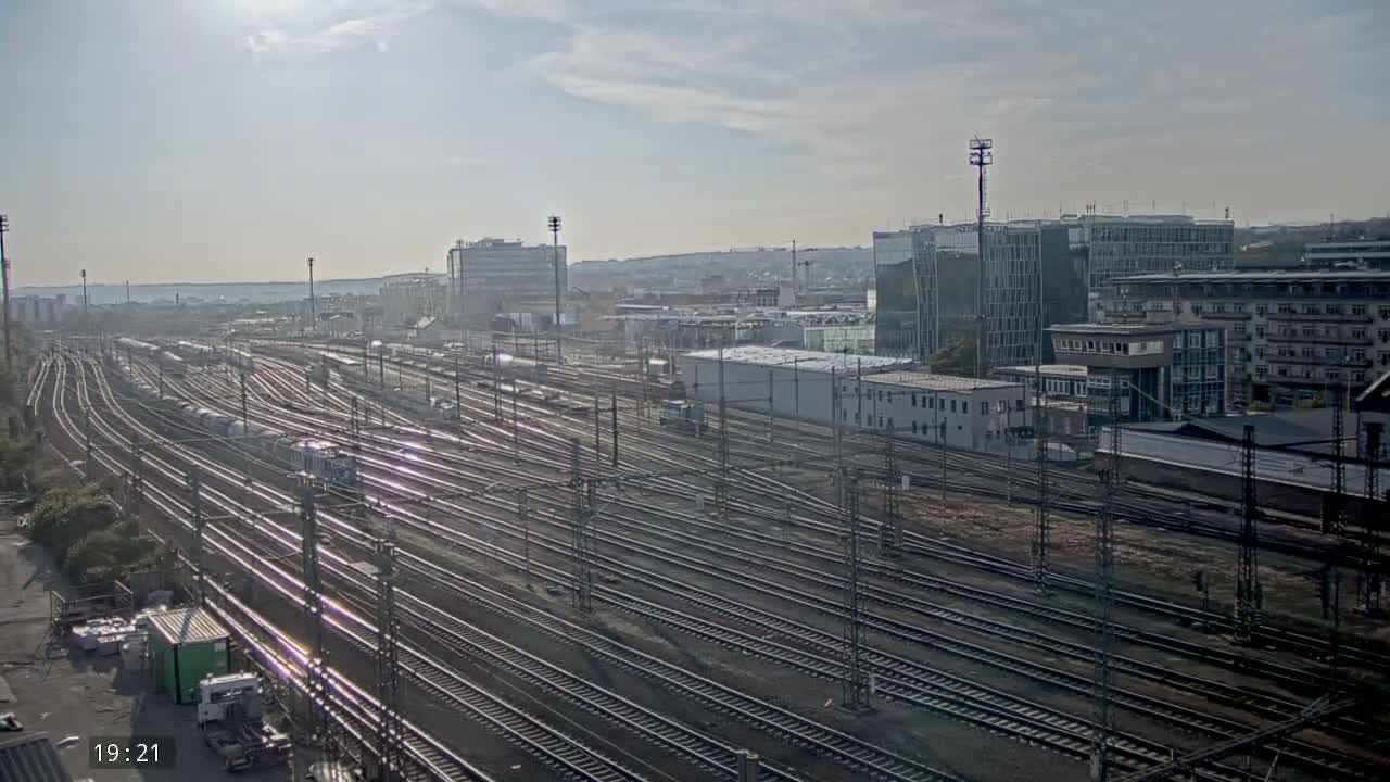 A large railway yard with numerous train tracks and parked freight cars stretches out before a cityscape under a mostly sunny sky.