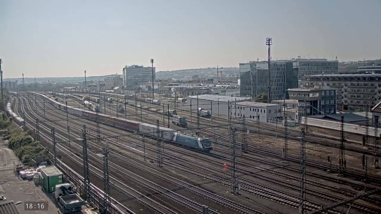 A wide view of a sprawling railway yard with numerous train cars and tracks under a clear, sunny sky, surrounded by industrial buildings and city structures.