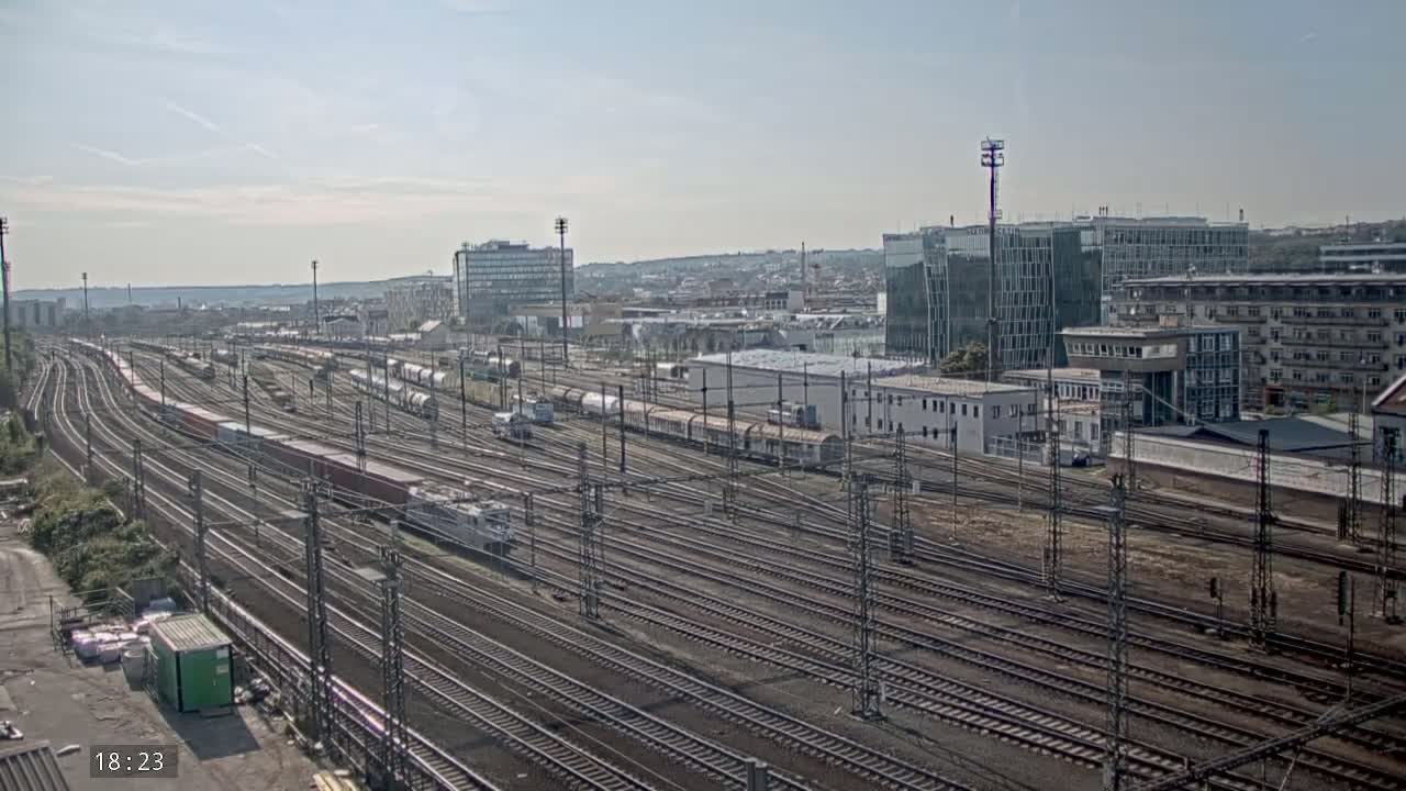 A sprawling railway yard with numerous train cars and tracks, under a mostly clear sky, is visible next to a city skyline on a sunny day.