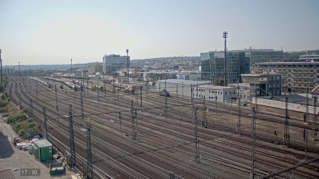 A wide shot on a sunny day shows numerous train tracks extending into the distance, with several trains and modern buildings visible in the background.