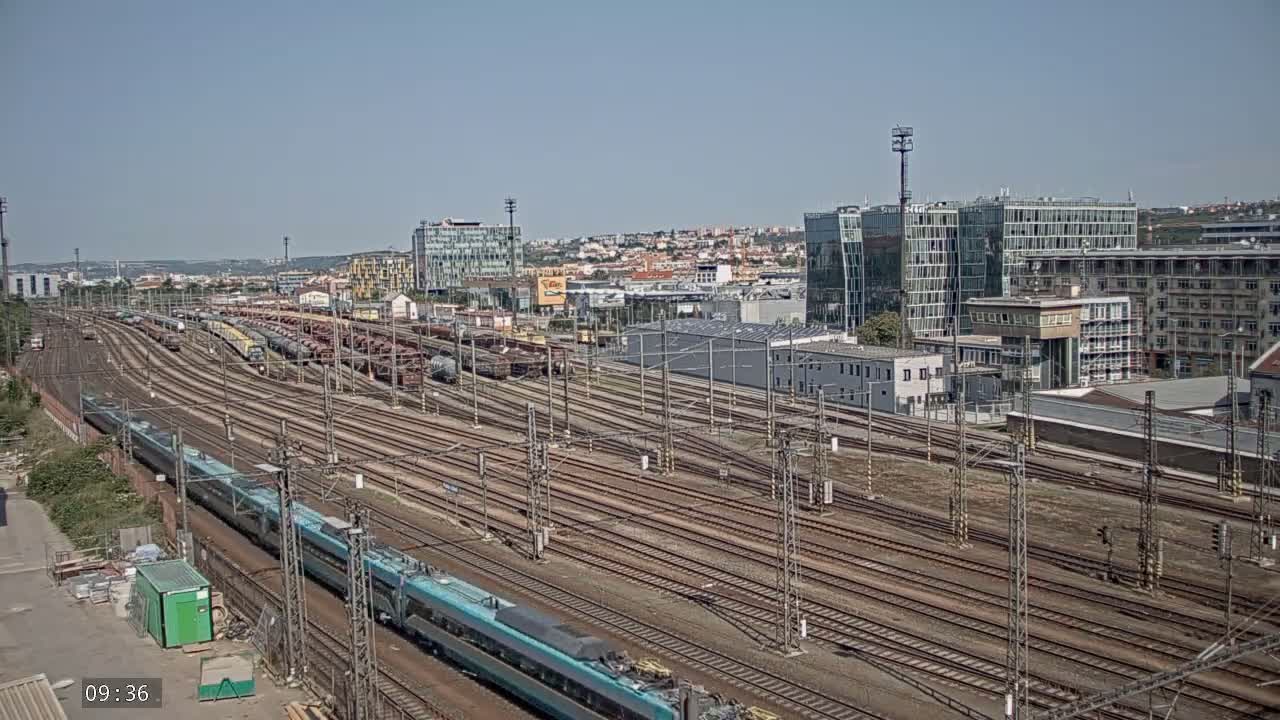 A long teal passenger train travels alongside numerous railway tracks, past a large freight train yard under a clear blue sky.