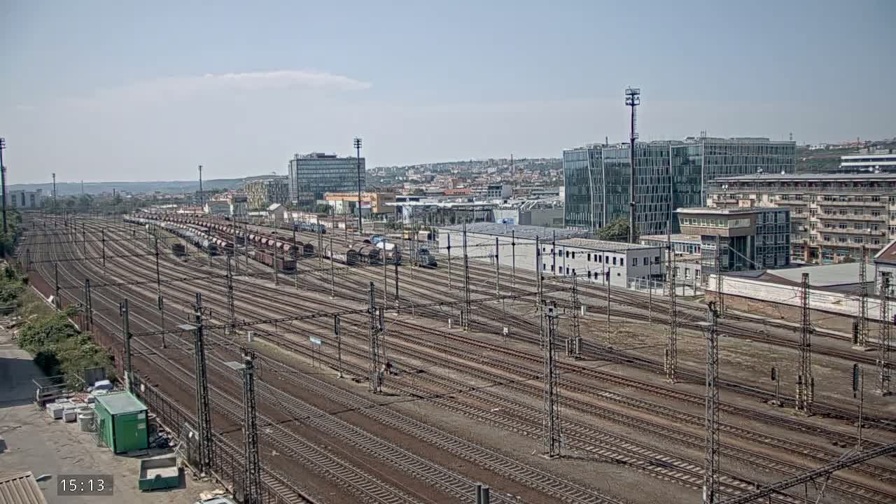 A large rail yard with numerous train cars and tracks extends into a cityscape under a mostly clear sky.