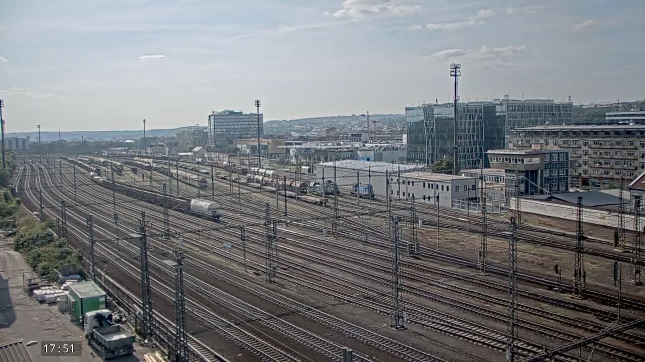A large rail yard with numerous train cars and tracks, under a partly cloudy sky, is situated near several buildings in a city.