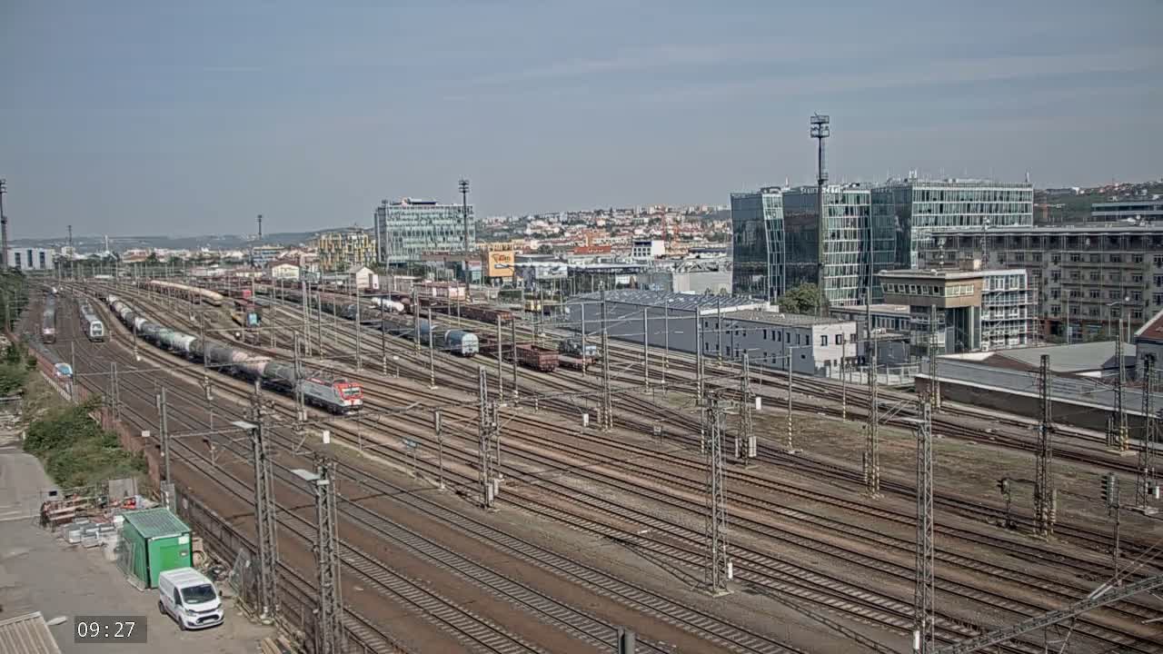 A sprawling railway yard with numerous train cars and tracks extends towards a cityscape under a clear sky.