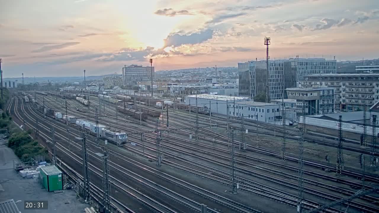 A sprawling train yard with numerous tracks and freight cars is visible at sunset under a partly cloudy sky.
