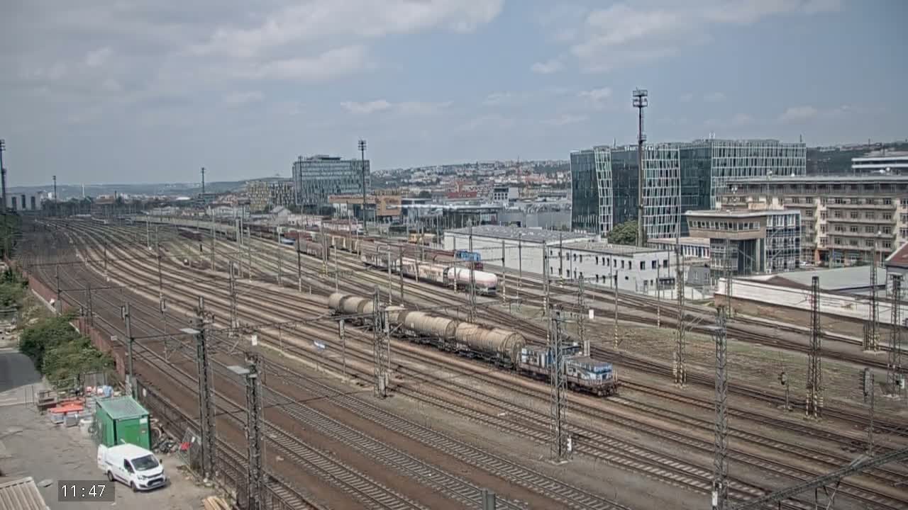 A sprawling railway yard with numerous tracks and trains under a partly cloudy sky, situated near a cityscape of modern and older buildings.