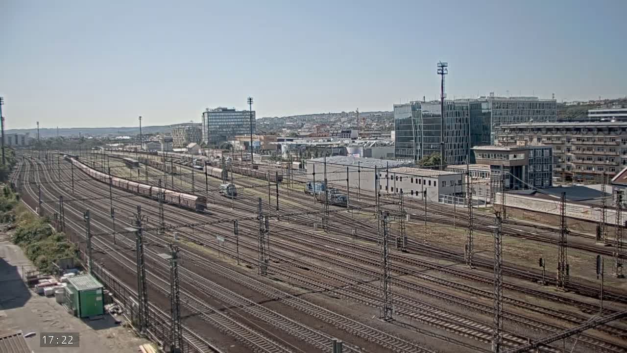 A long freight train sits on a sprawling network of train tracks under a clear, sunny sky, with modern buildings visible in the background.