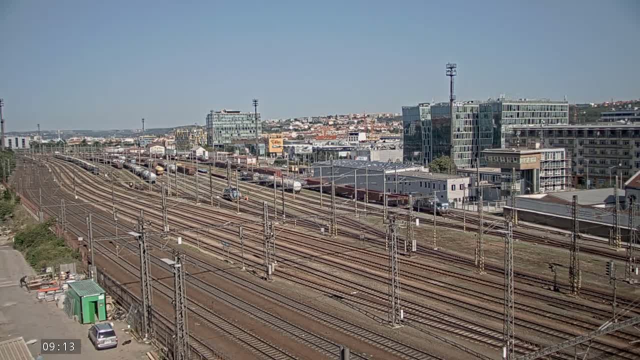 A vast railway yard with numerous train tracks and parked freight cars extends beneath a clear, sunny sky toward a cityscape in the distance.