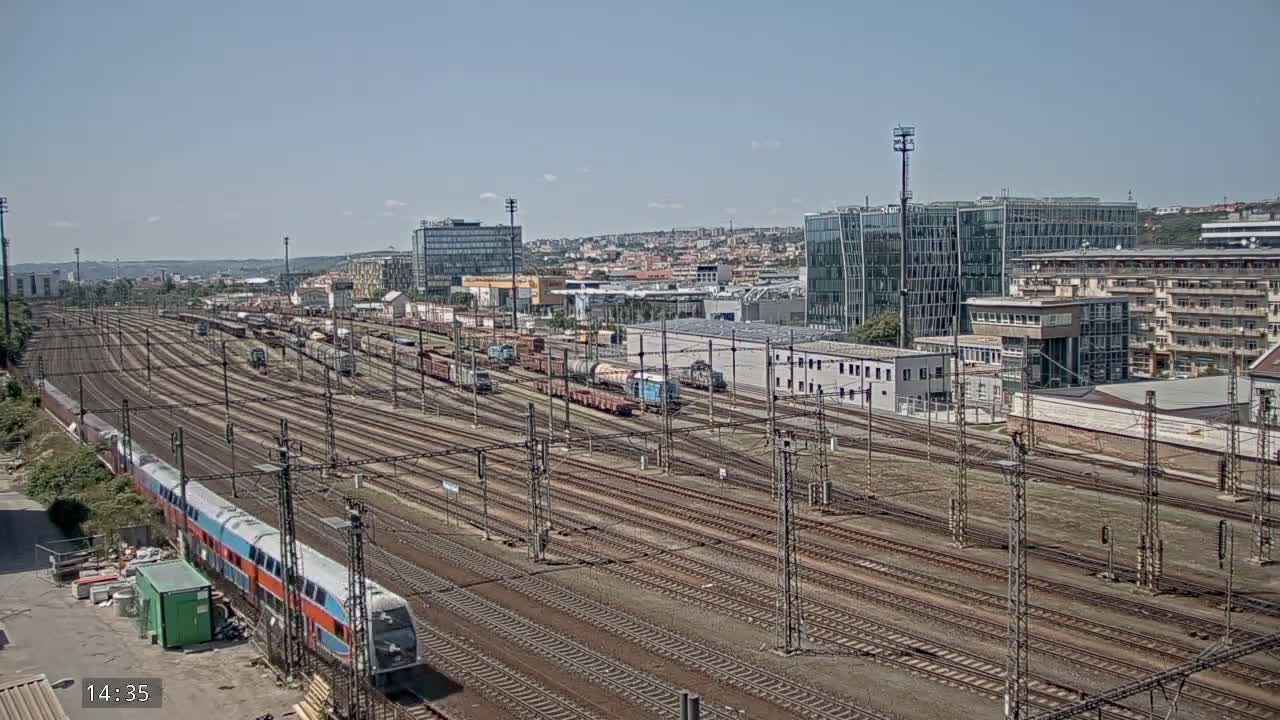 A long train and numerous freight cars are situated on a sprawling railway network under a clear, sunny sky, with a cityscape visible in the background.
