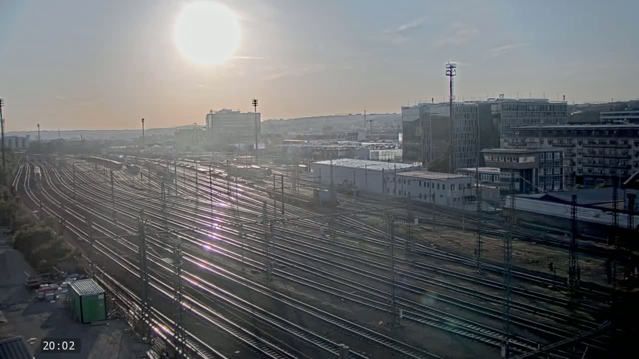 A hazy sunrise illuminates a vast network of train tracks surrounded by industrial buildings and a cityscape in the background.