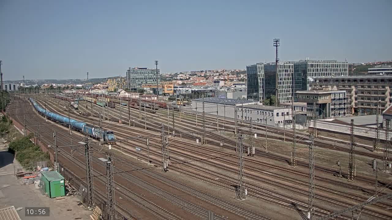 A long blue train travels along numerous train tracks in a sunny urban area with several buildings in the background.