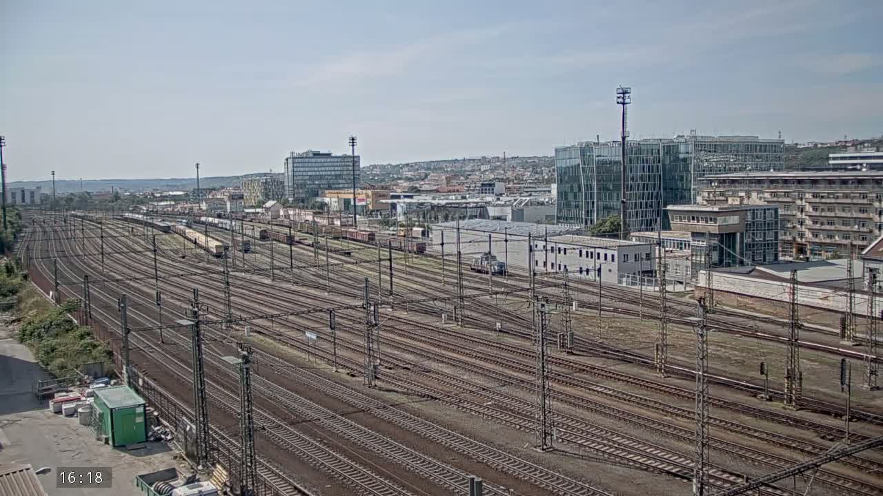 A vast network of train tracks extends into the distance, under a clear, sunny sky, past various buildings and parked trains.