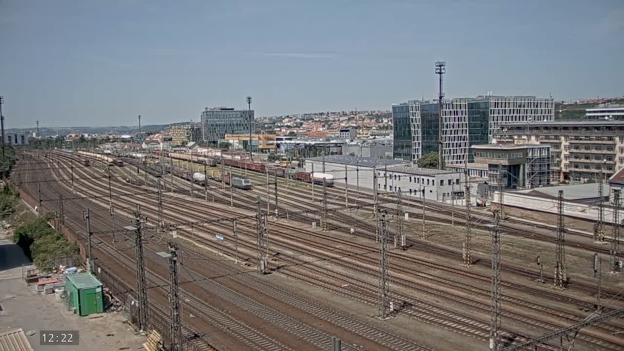 A wide shot on a sunny day shows numerous railway tracks extending into the distance, alongside freight trains and various buildings, including modern high-rises and older structures.