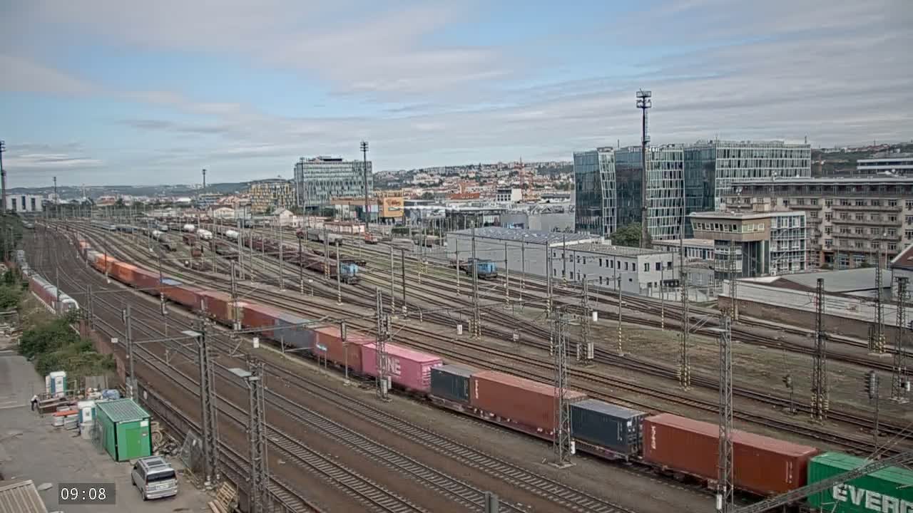 A large railway yard with numerous trains and cargo containers is shown under a partly cloudy sky.