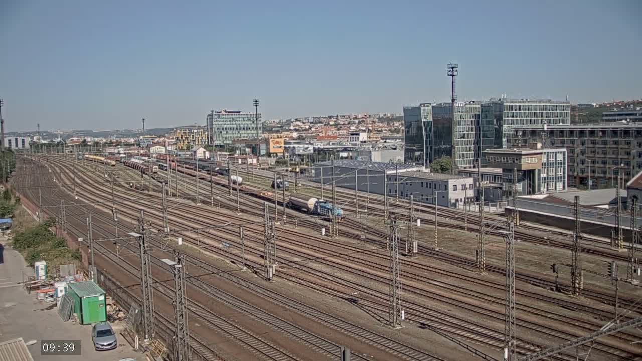 A vast railway yard with numerous tracks extends into the distance under a clear blue sky, surrounded by modern buildings and a cityscape.