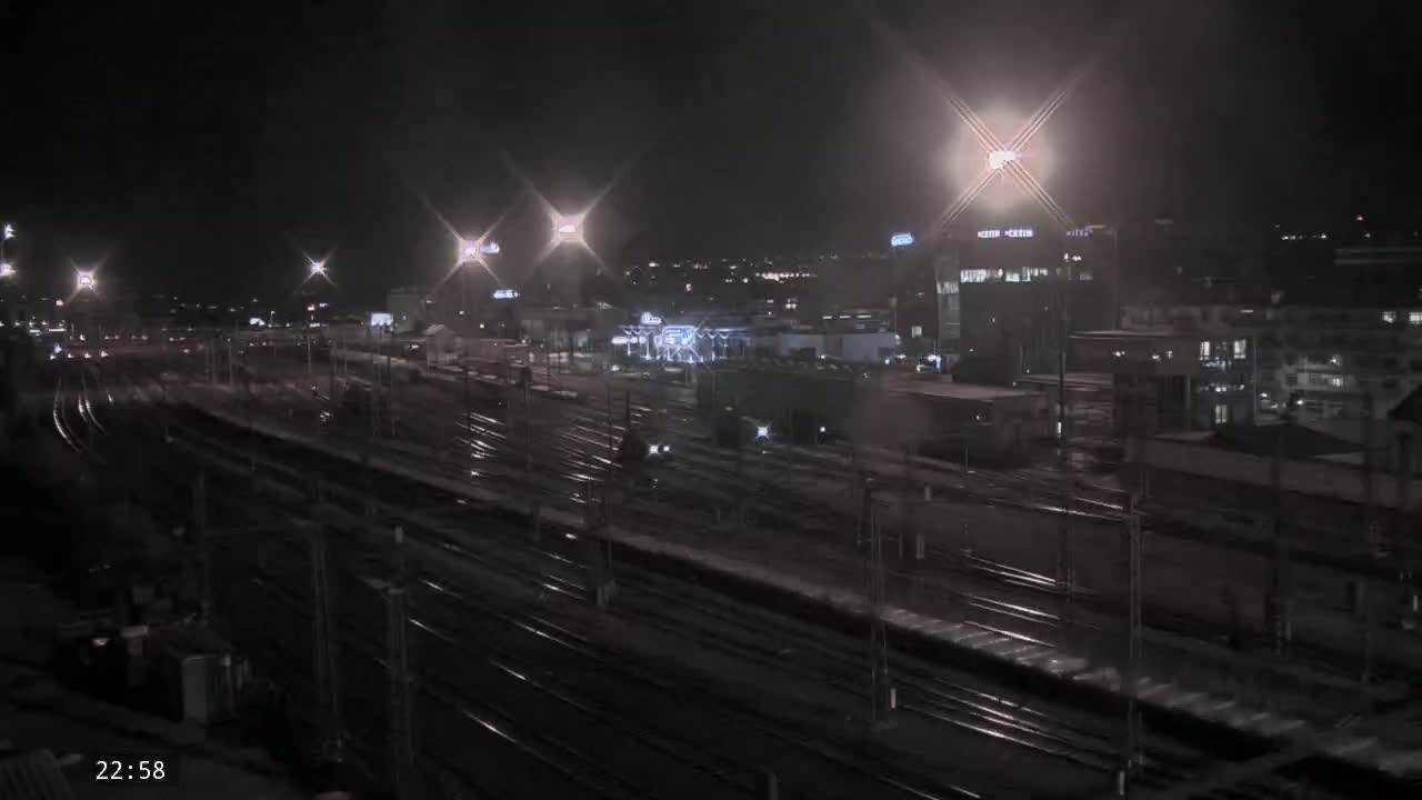 A nighttime view of a train yard with several tracks and buildings in the background, possibly under light snow or rain.