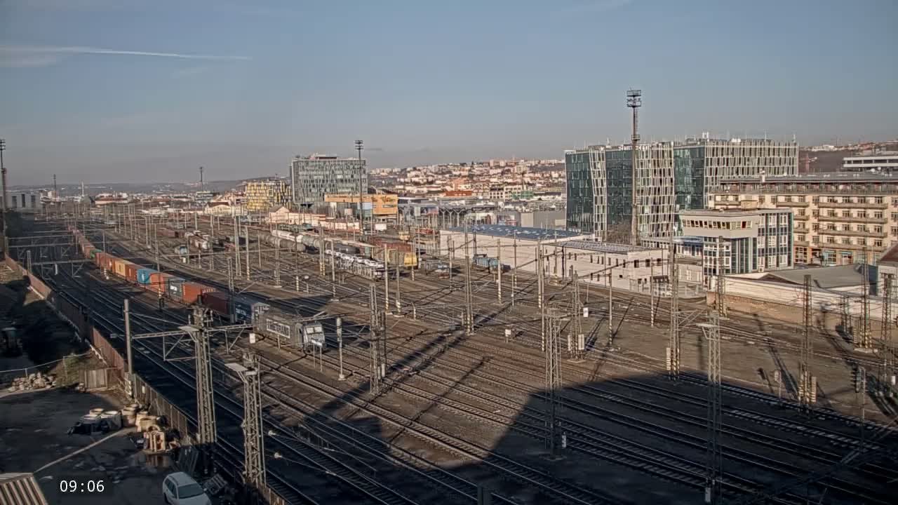 An elevated view reveals an expansive train yard with multiple tracks, several trains, and overhead power lines, set against a backdrop of a city with various modern and older buildings, under a clear, sunny sky.
