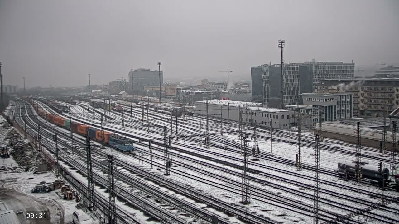 A sprawling snow-covered railway yard, bustling with numerous tracks and freight trains, leads to a distant urban skyline under a dreary, overcast winter sky.