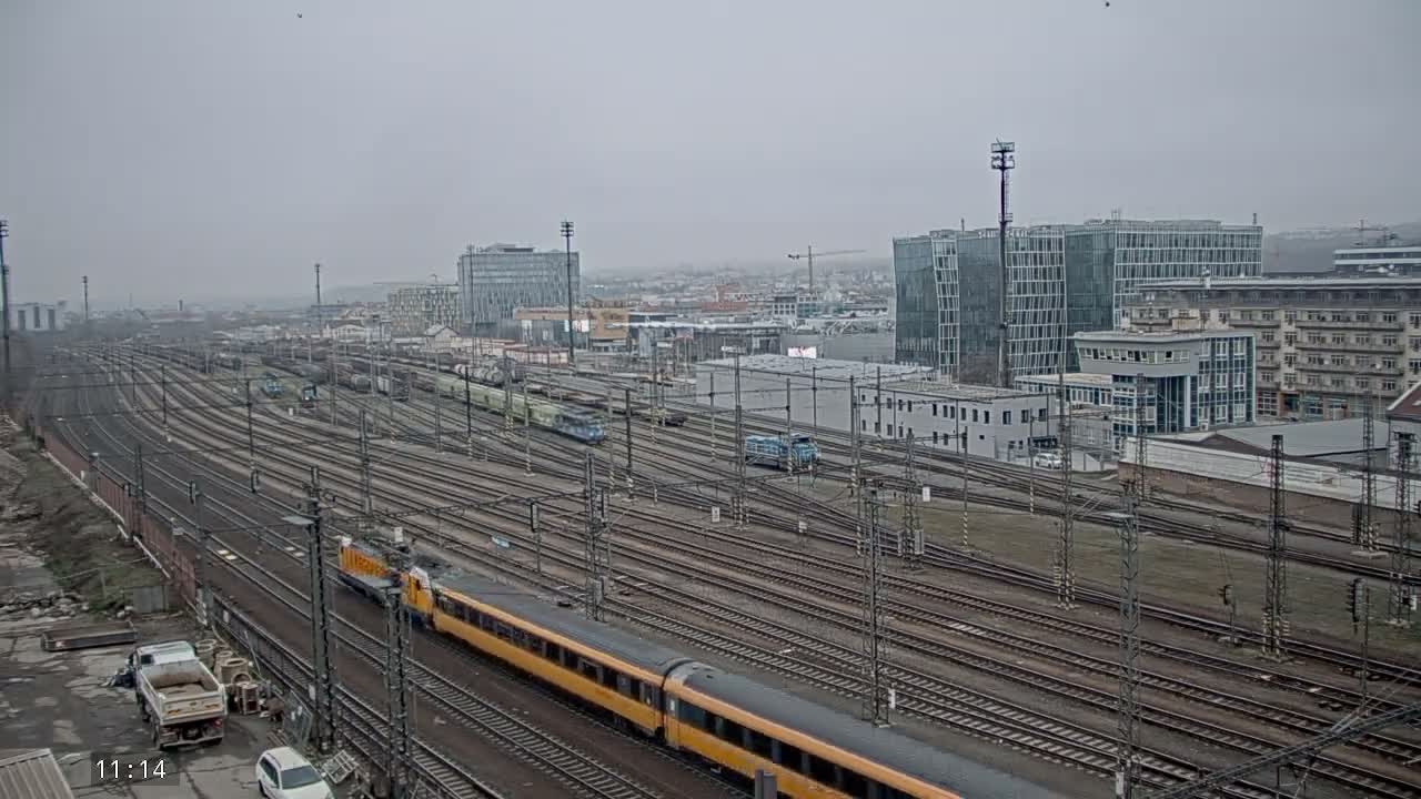 Under an overcast sky, a large urban railway junction is filled with numerous tracks and multiple trains, including a prominent yellow passenger train in motion, all set against a backdrop of diverse city buildings.