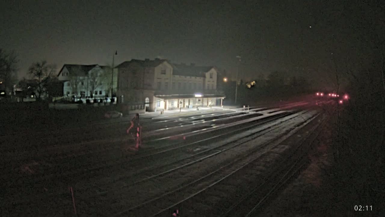 A nighttime view of a train station and tracks, with a person walking near the station and faintly lit buildings in the background.