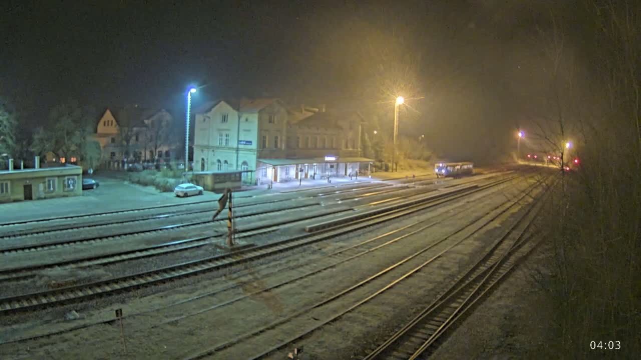 A nighttime view of a train station with multiple train tracks covered in a light dusting of snow or frost, under hazy, dimly lit conditions.
