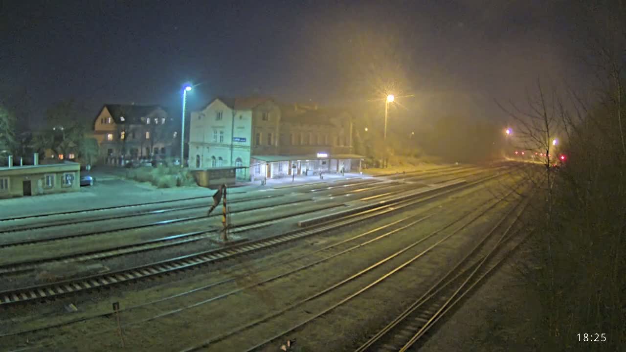 A nighttime view of a train station with multiple tracks extending into a foggy distance, illuminated by streetlights.