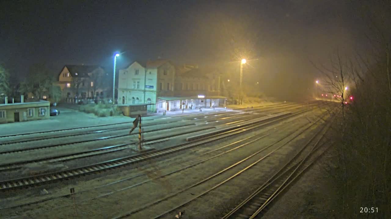 A nighttime view of a train station with several tracks extending into a foggy distance, illuminated by streetlights.