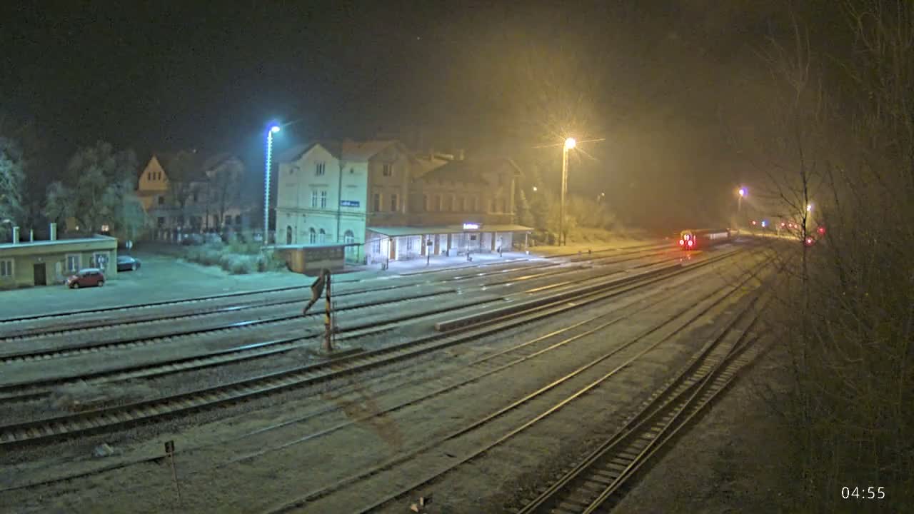 A train station at night under foggy conditions, with a train departing and several tracks covered in a light layer of snow or frost.