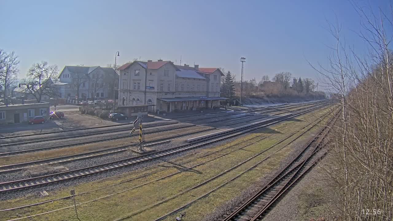 A train station with a long platform and several tracks is visible on a sunny day with patches of snow and bare trees in the background.