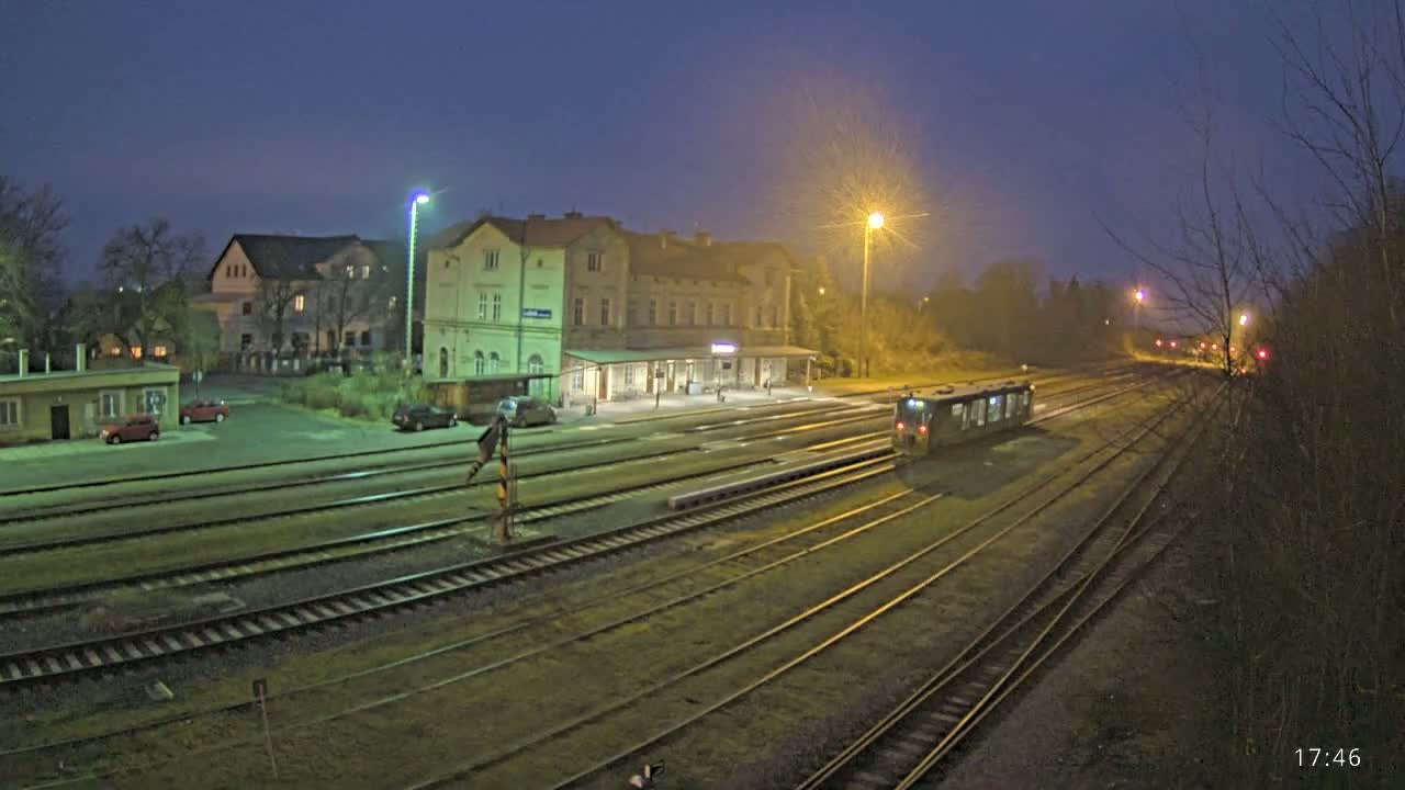A train sits in a station at night, under hazy streetlights, next to a long building with several cars parked nearby.
