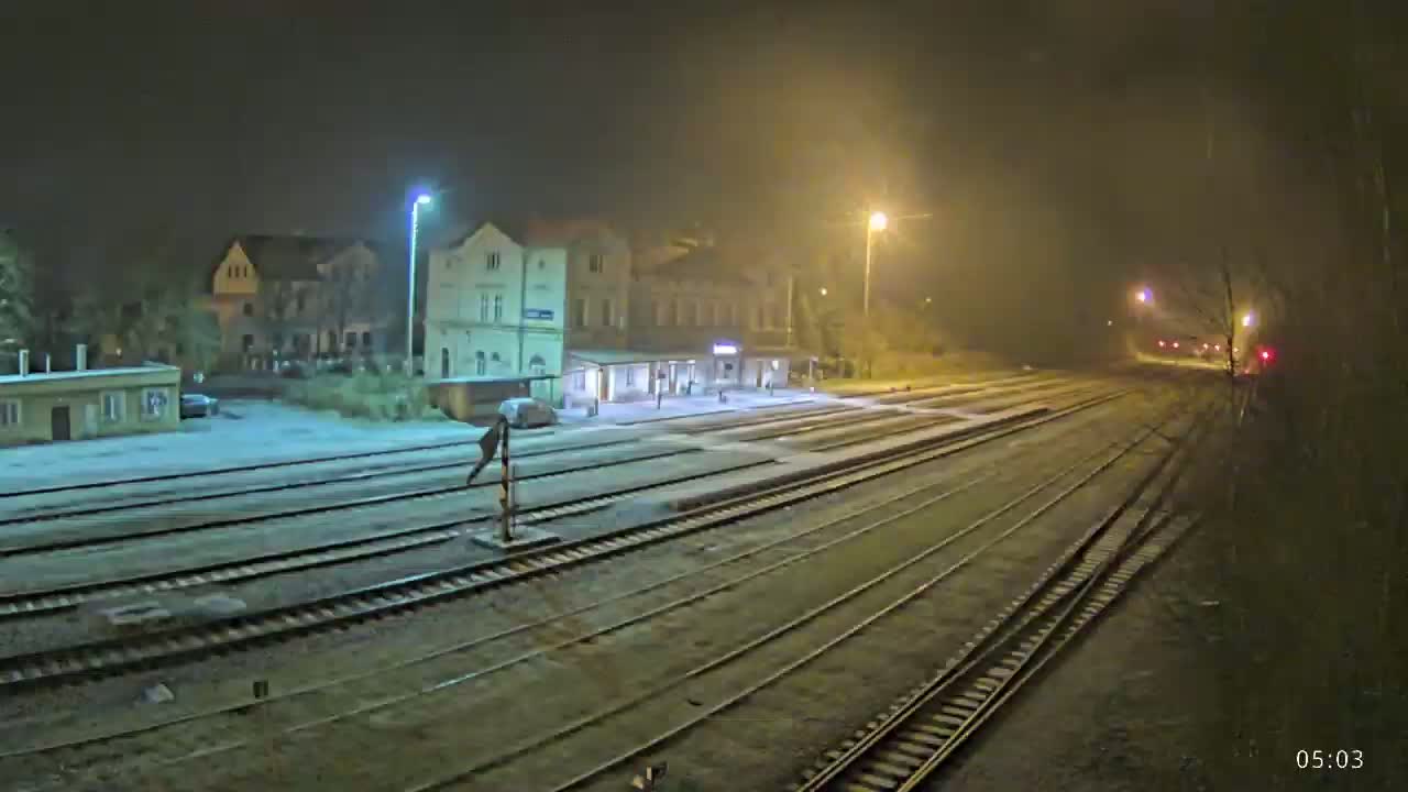 A snow-covered train station at night, illuminated by streetlights, with multiple train tracks extending into the distance.