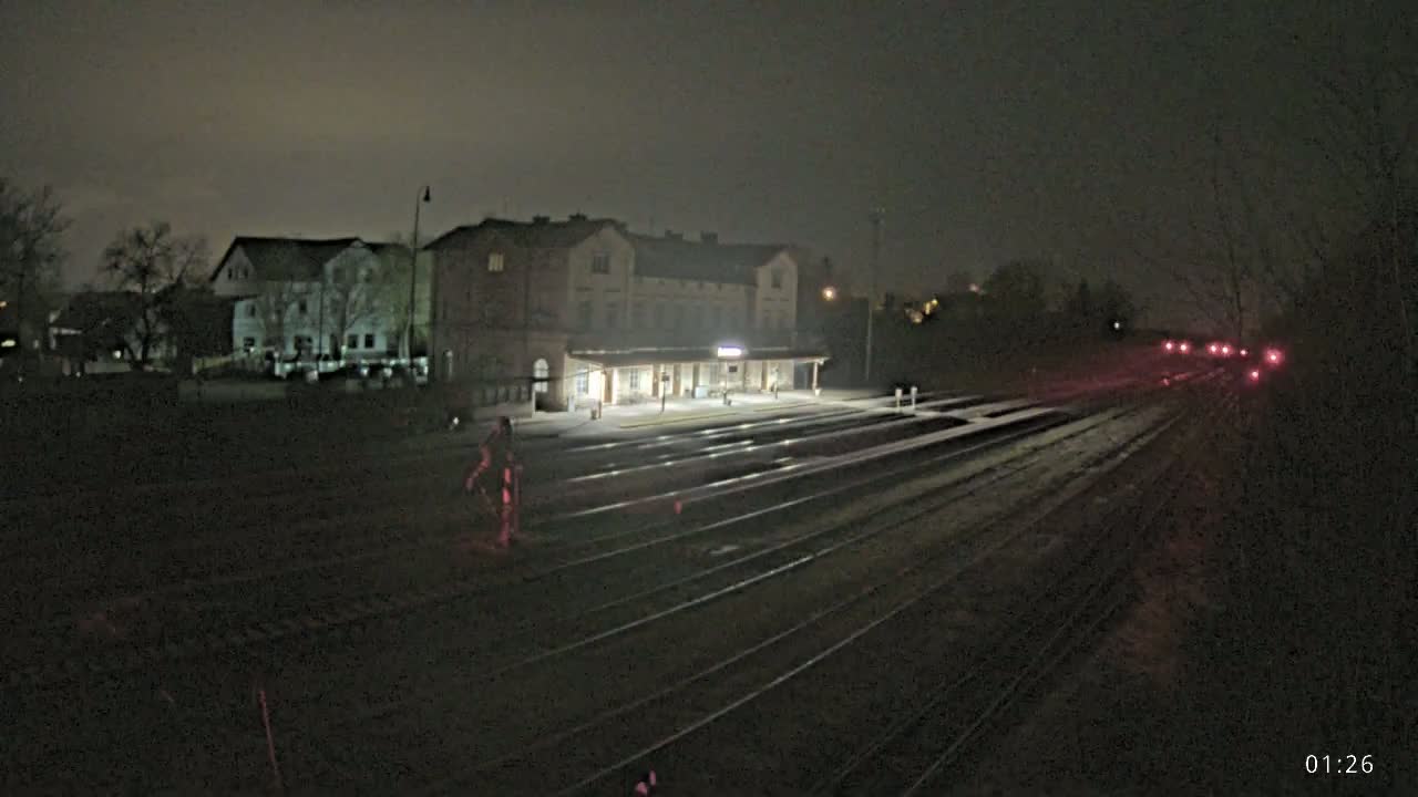 A train station at night, under a dark sky, with several sets of train tracks extending into the distance and a few distant lights visible.