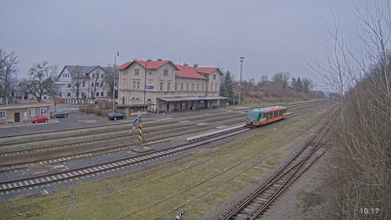 A light-green train is arriving at a train station on an overcast day, with several cars parked nearby.