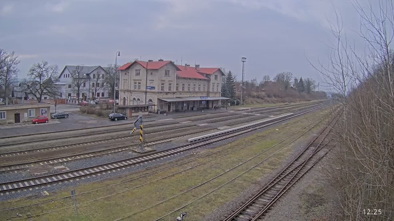 A train station with a long platform and several tracks is visible under an overcast sky.