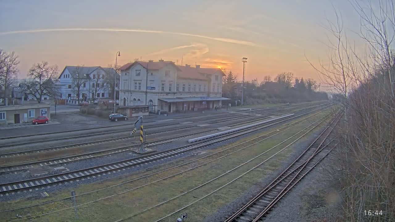 A train station with multiple tracks is seen at sunset, with a few cars parked nearby and bare trees in the background.