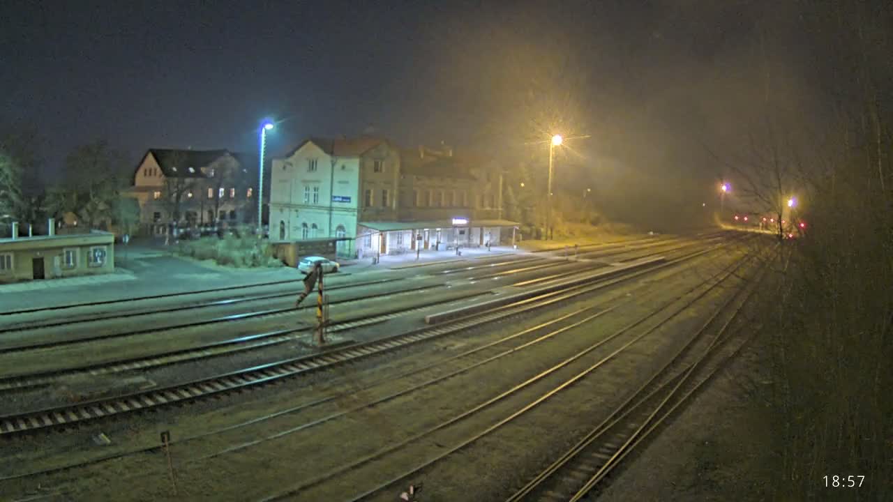 A nighttime view of a train station with multiple tracks extending into the foggy distance, illuminated by streetlights.