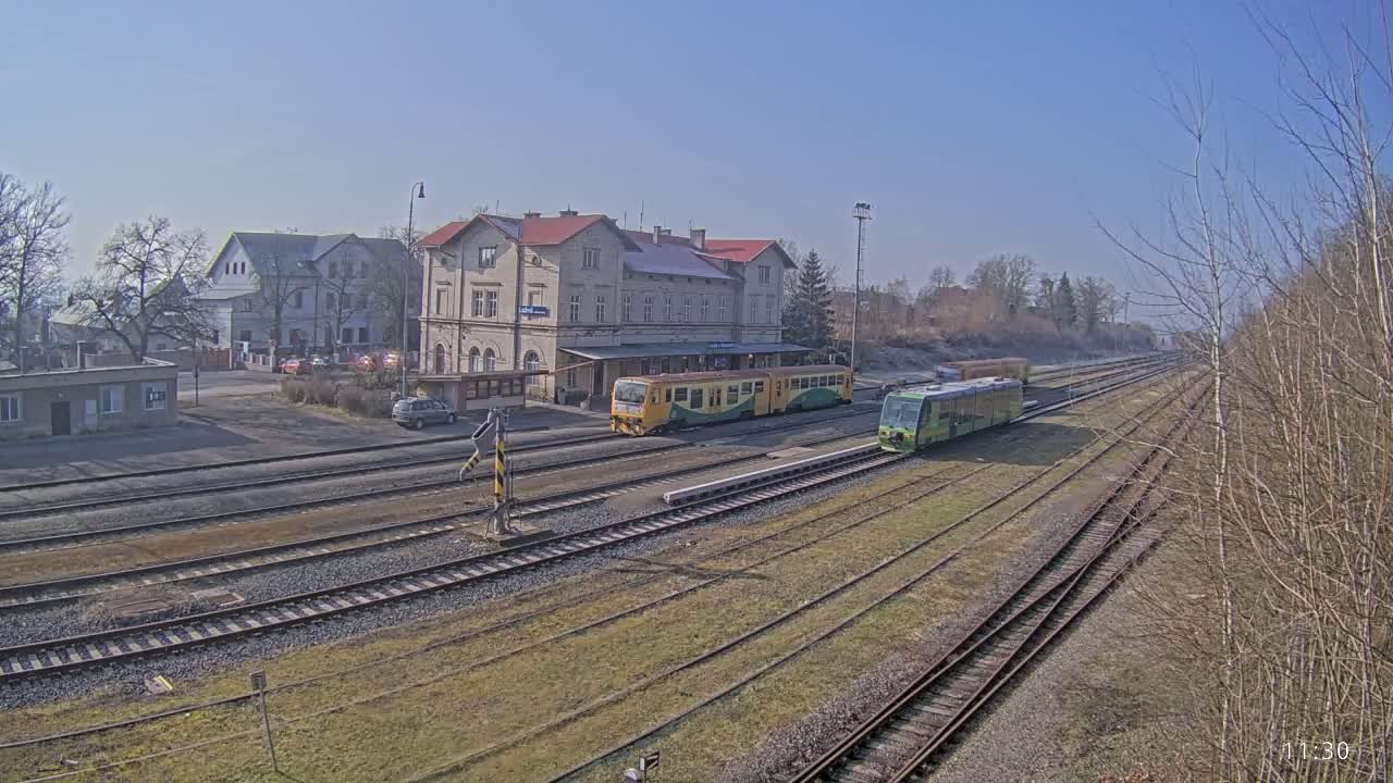 Two trains are parked at a train station on a sunny day, next to a long building with a red roof.