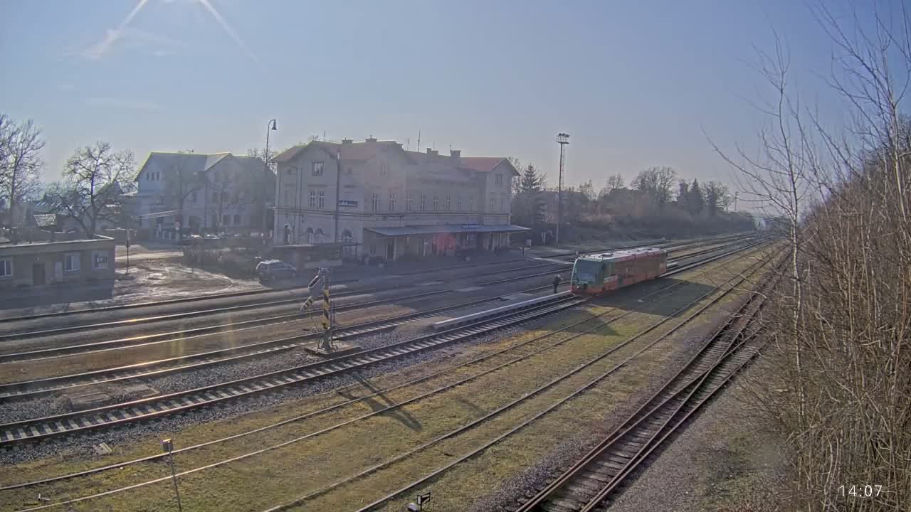 A train sits at a station on a sunny day, with a large building behind it and multiple train tracks extending into the distance.