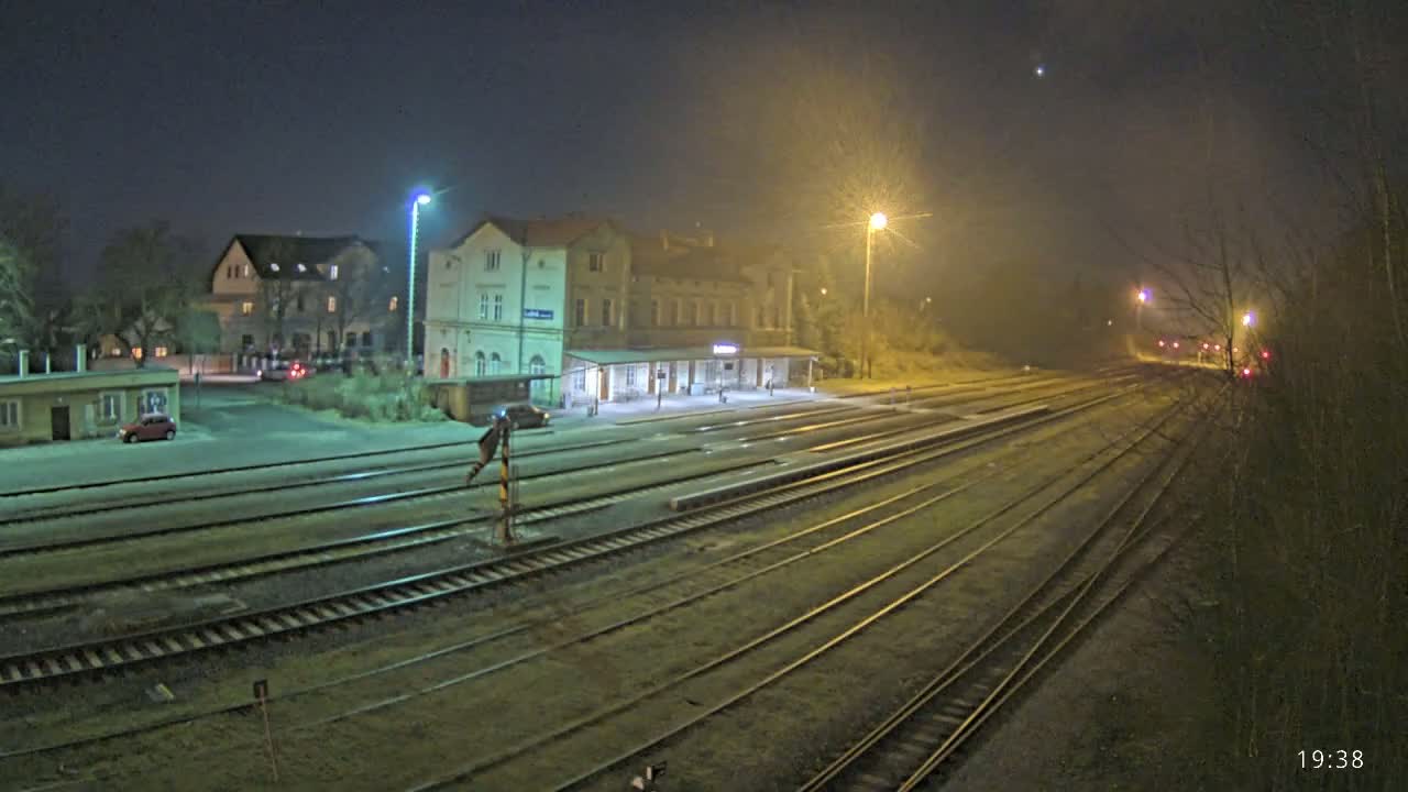 A nighttime view of a train station with a long expanse of train tracks extending into a foggy distance, illuminated by streetlights.
