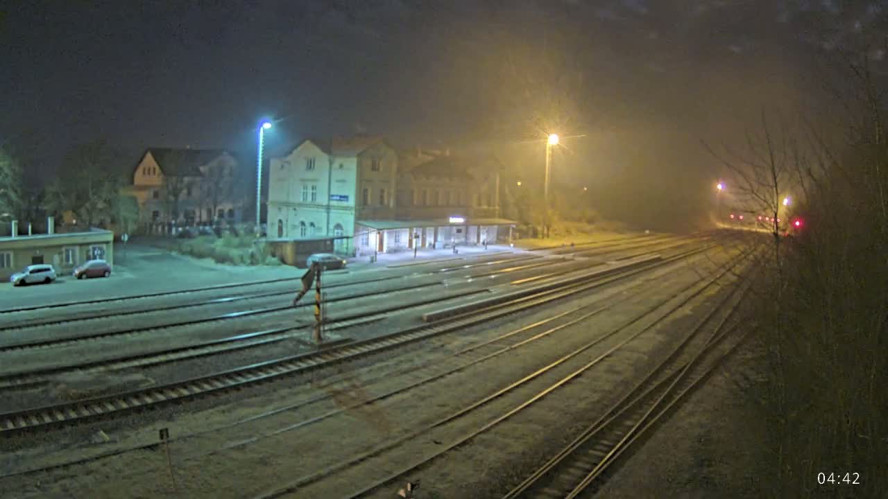 A train station at night in foggy conditions, with several tracks extending into the distance, and a few cars parked nearby.