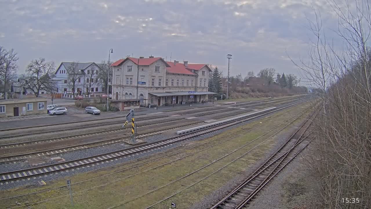 A long view of a train station with a red-roofed building, several railway tracks, and a few cars parked nearby on an overcast day.