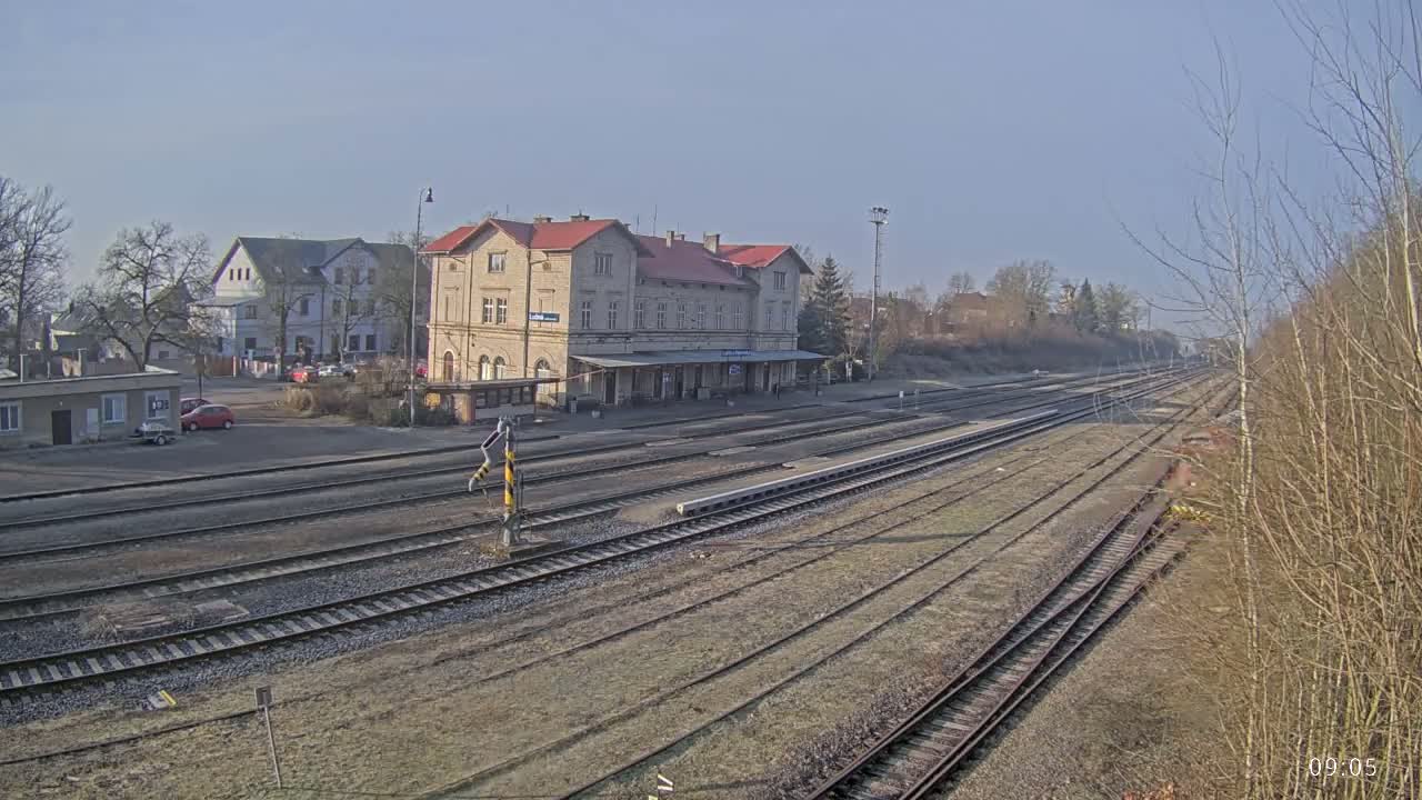 A long view of a train station on a sunny day shows multiple train tracks extending into the distance, with a large stone building at the center and other buildings and cars visible in the background.