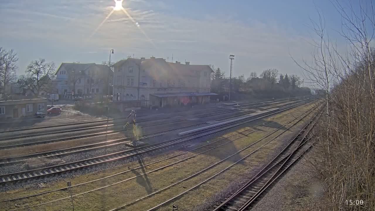 A sunny day reveals a train station with multiple tracks extending into the distance, surrounded by low buildings and bare trees.