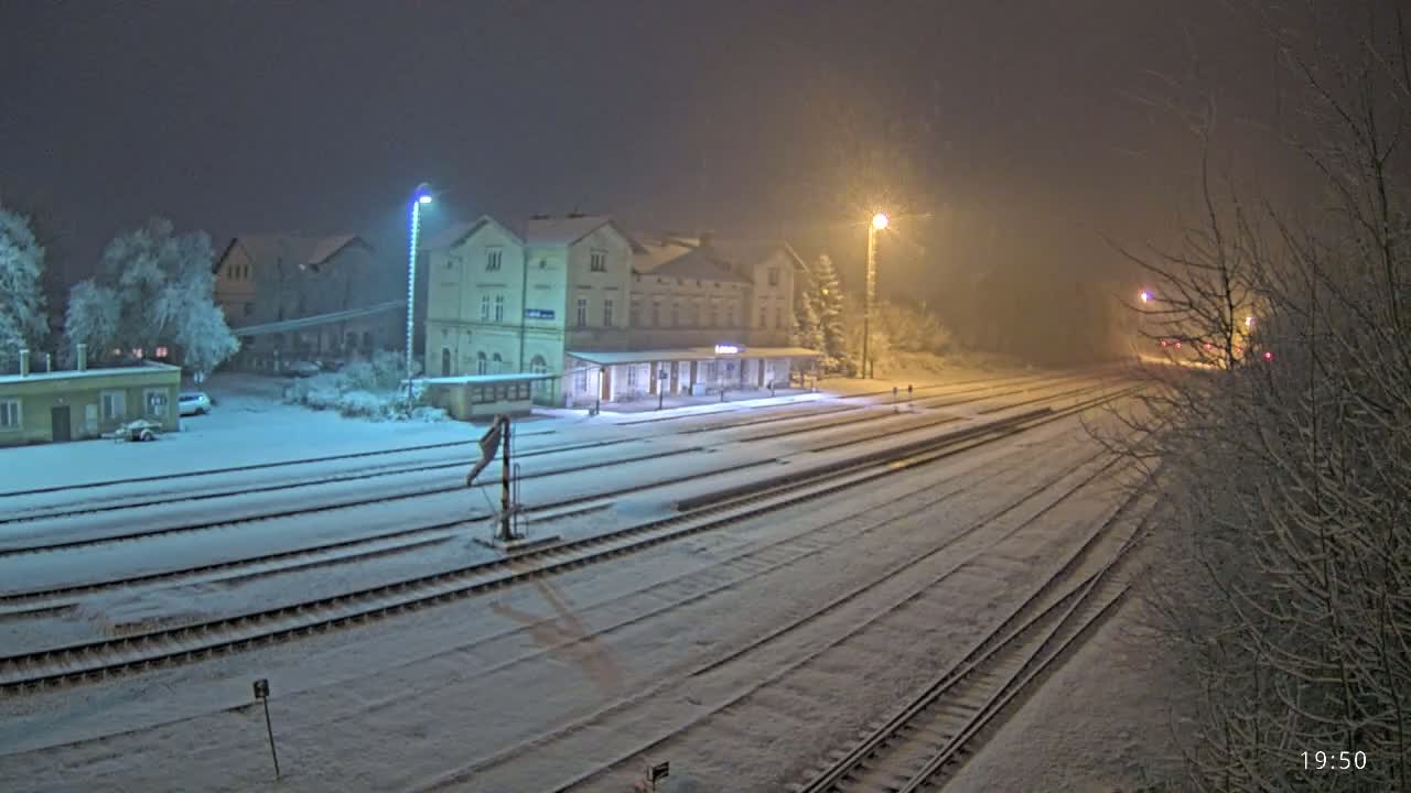 A snow-covered train station at night, illuminated by streetlights, with multiple train tracks extending into the hazy distance.