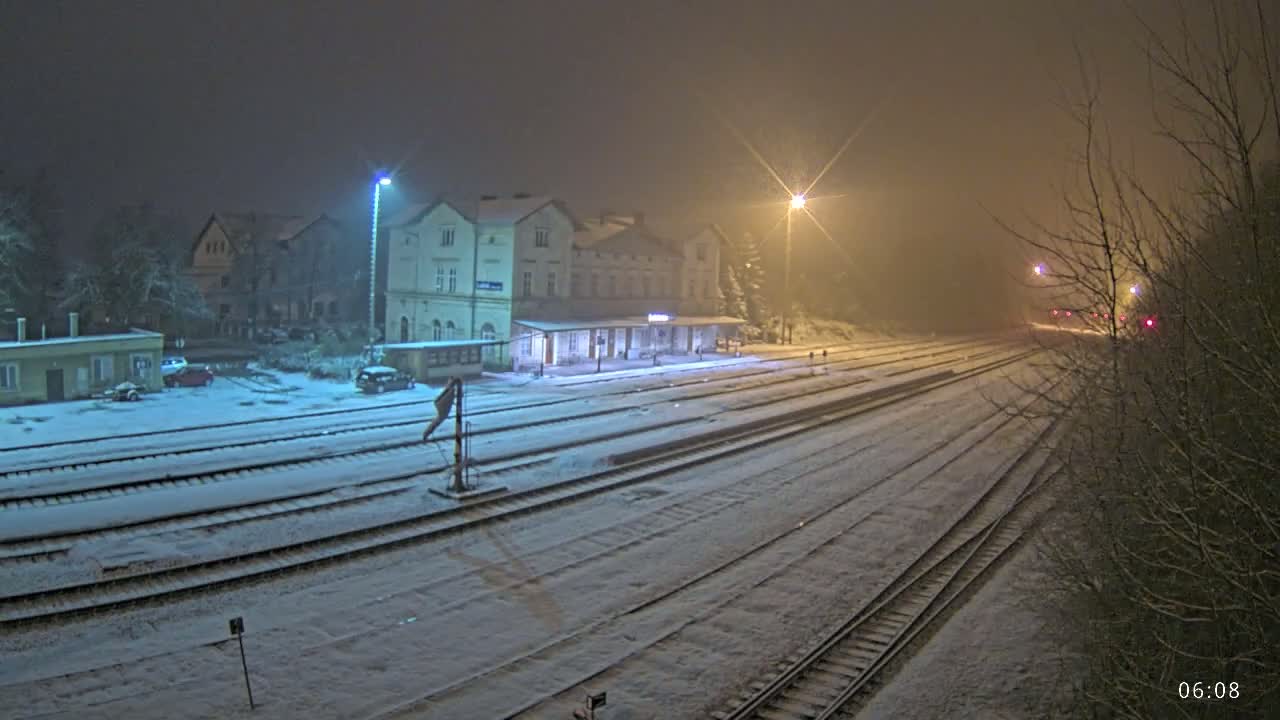A snow-covered train station at night is illuminated by streetlights, with several train tracks extending into the foggy distance.