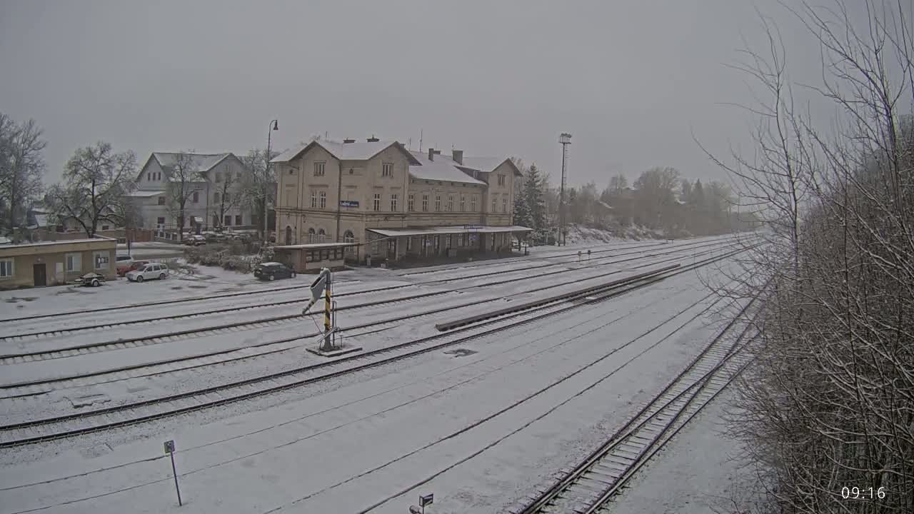 A snow-covered train station and several tracks are visible in the lightly snowing weather.