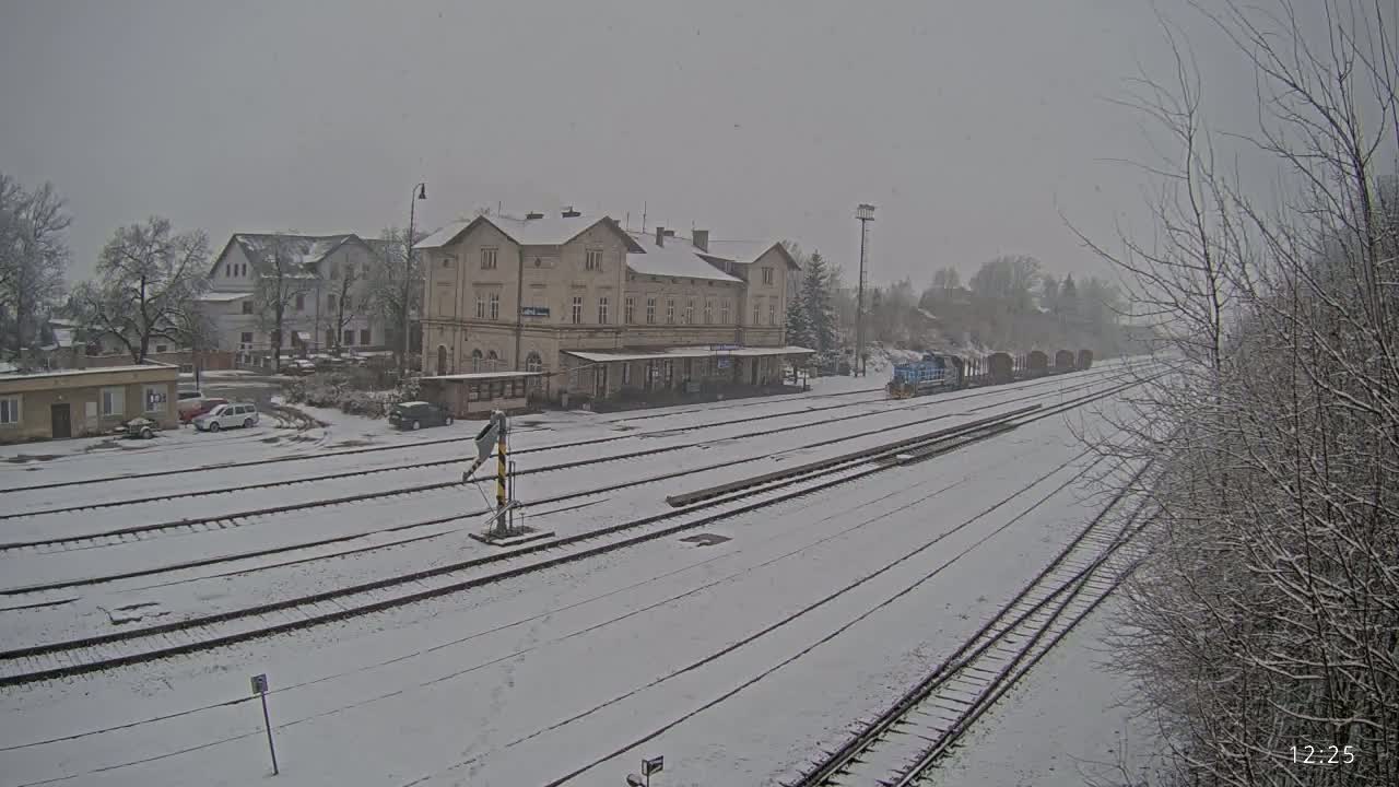 A snow-covered train station with a long building and several train tracks, and a freight train in the distance under a light snowfall.