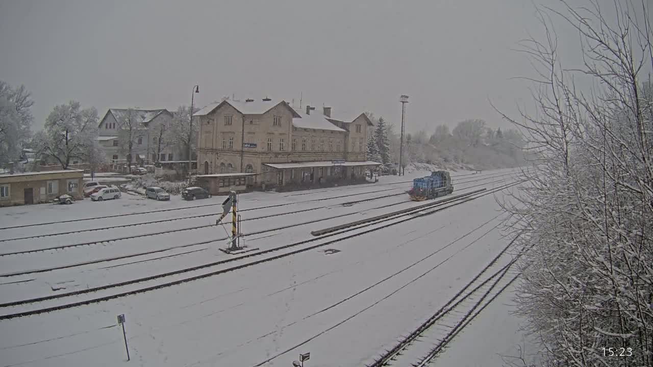 A snow-covered train station with a lone train and several cars parked nearby, under a gray, snowy sky.