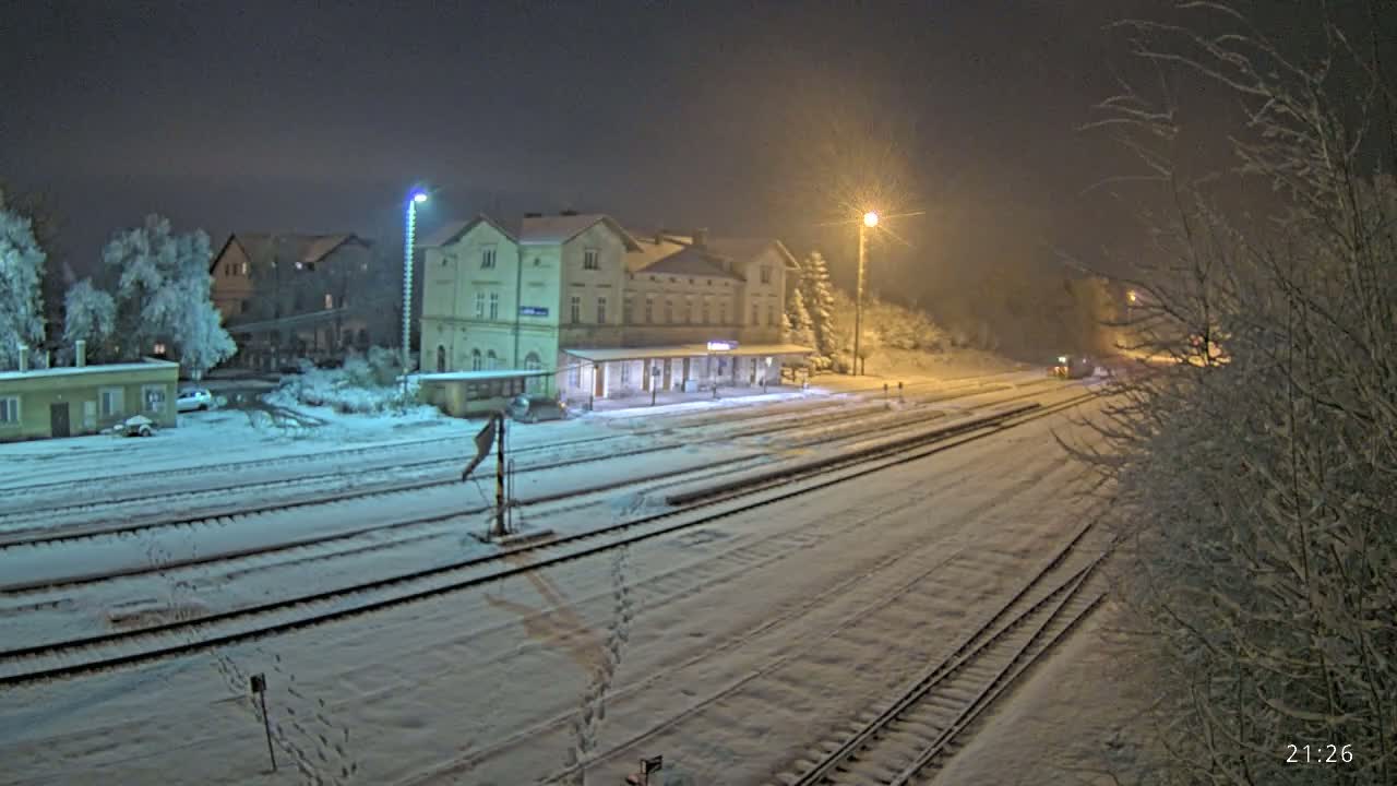 A snow-covered train station at night, illuminated by streetlights, with several buildings and tracks visible.